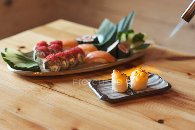 From above plate with assorted sushi rolls being burned with torch by cropped unrecognizable cook served on table in Japanese restaurant — Stock Photo
