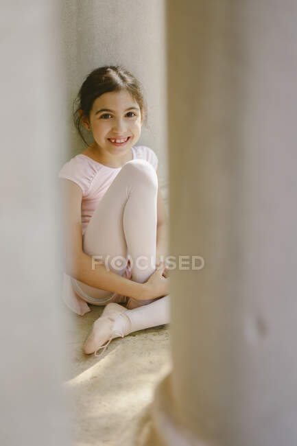 Positive girl in ballet uniform sitting near stone column in park and looking at camera — Stock Photo
