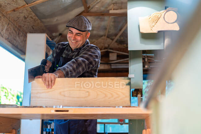 Happy carpenter using sharp saw while cutting lumber plank at workbench in professional workshop — Stock Photo