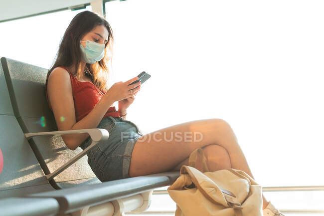 Женщина-туристка в защитной маске сидит в зале вылета аэропорта и ждет полета во время эпидемии коронавируса при использовании смартфона — стоковое фото