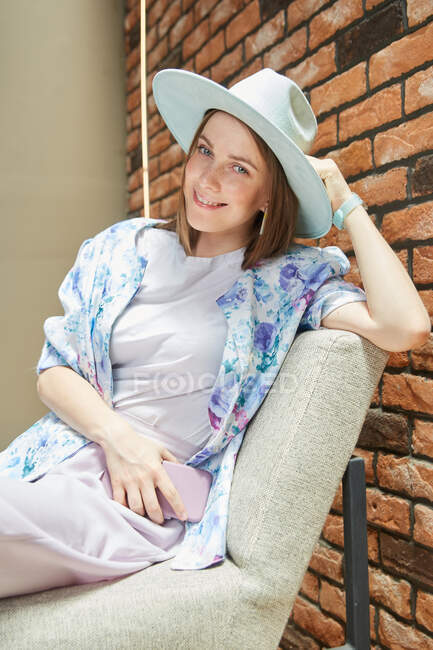 Молодая женщина в блузке с цветочным орнаментом сидит на стуле со смартфоном, глядя в камеру — стоковое фото