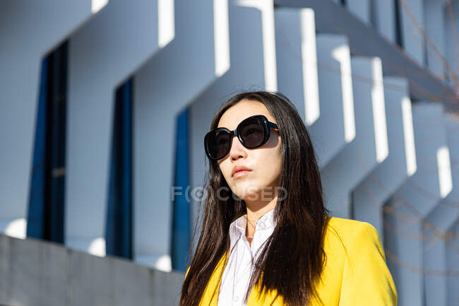 Mujer de negocios asiática con abrigo amarillo caminando por la calle con el edificio en el fondo - foto de stock