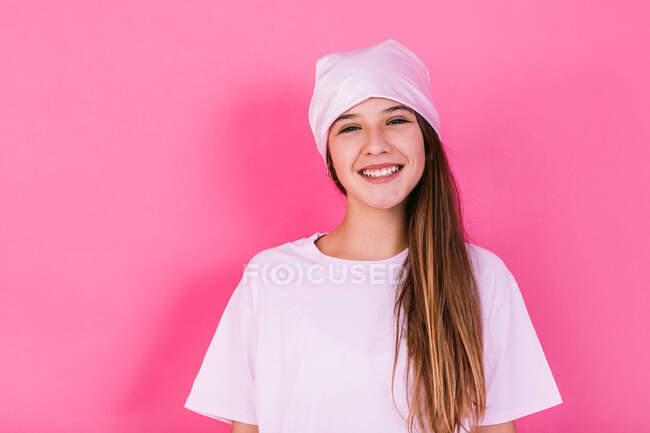 Adolescente feliz do sexo feminino com cabelo castanho e lenço de cabeça representando a consciência do câncer olhando para a câmera no fundo rosa — Fotografia de Stock