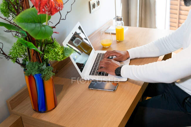 High angle of crop anonymous ethnic businessman typing on netbook sitting at table in hotel room during business trip — Stock Photo