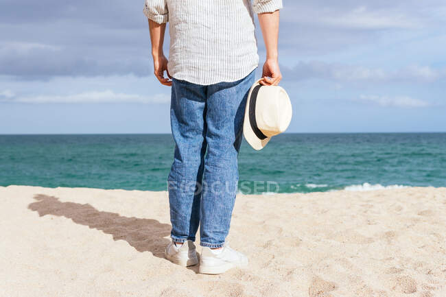 Back view of cropped unrecognizable male in trendy casual clothes and hat walking alone on sandy beach towards waving sea while spending summer holidays on seashore — Stock Photo