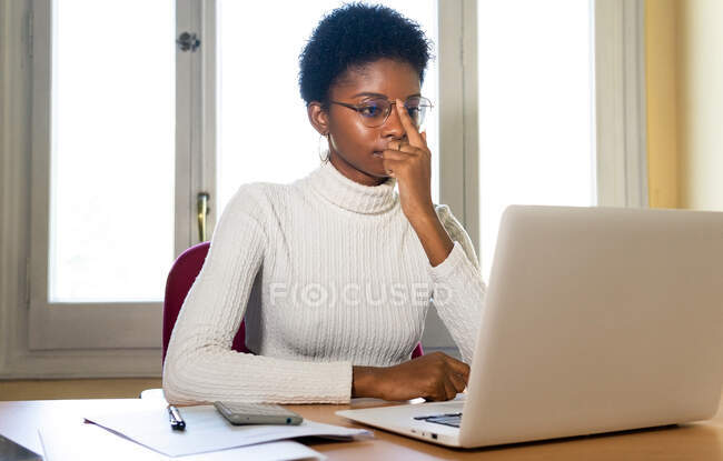 Сконцентрована молода афроамериканська жінка в повсякденному одязі і окулярах дивлячись на екран ноутбука і читання професійної інформації під час роботи за столом в офісі — стокове фото