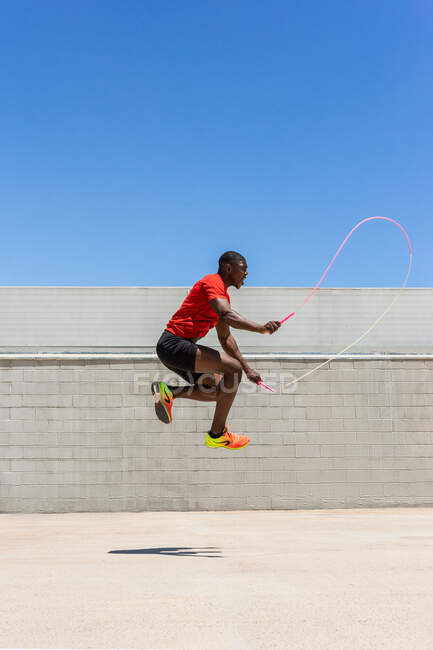 Vista lateral do atleta masculino afro-americano enérgico pulando corda acima do solo enquanto faz exercícios durante o treinamento no verão — Fotografia de Stock