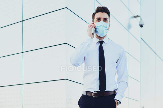 Stylish male executive in formal clothes and medical mask talking on cellphone while looking away in town — Stock Photo