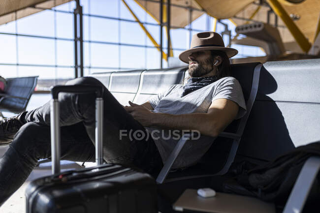 The guy with a hat at the airport in the waiting room sitting waiting for his flight, with wireless headphones to listen to music, sleeps and with his hat covers his eyes — Stock Photo