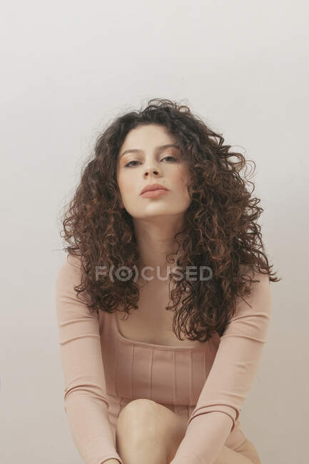 Dolce donna sognante con i capelli ricci e in body seduta sul pavimento contro parete bianca in studio — Foto stock