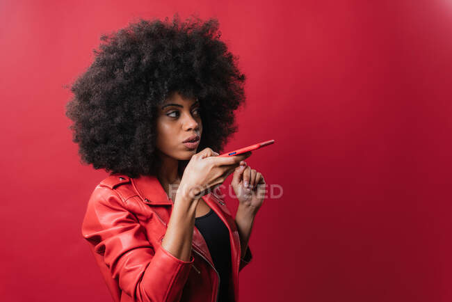 Стильная афроамериканка с прической в стиле афро записывает аудиосообщение на мобильный телефон на красном фоне в студии — стоковое фото