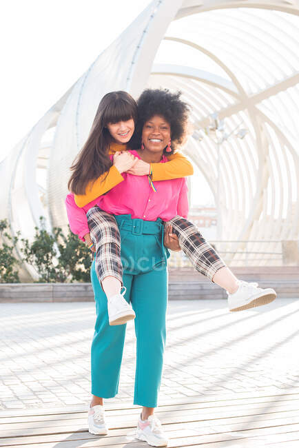 Smiling black woman piggybacking delighted lesbian girlfriend while having fun in city at weekend and looking at camera — Stock Photo