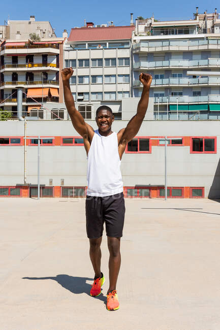Positive African American male athlete in sportswear standing with raised arms on sports ground and celebrating achievement while looking at camera — Stock Photo
