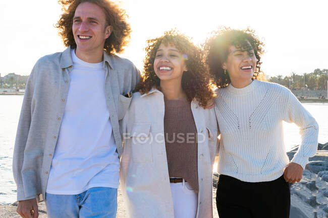 Group of young diverse women and man with curly hair embracing each other while walking on shoreline of cityscape in back lit — Stock Photo