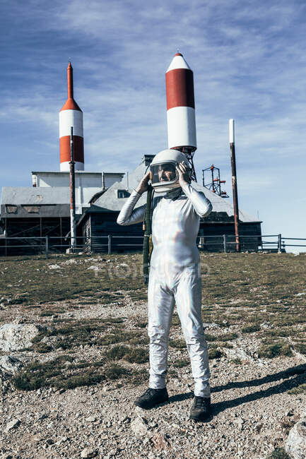 Homme au corps entier en combinaison spatiale debout sur un sol rocheux contre une clôture métallique et des antennes en forme de fusée rayées le jour ensoleillé — Photo de stock