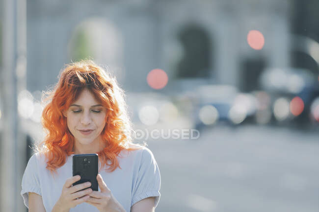 Весела руда жінка на вулиці і повідомлення в соціальних мережах на мобільному телефоні — стокове фото