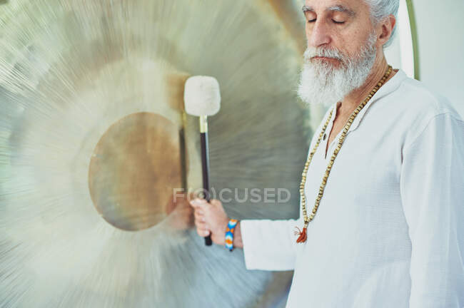 Vista laterale del maschio adulto in abiti bianchi con martello che gioca a gong sospeso durante la pratica spirituale — Foto stock