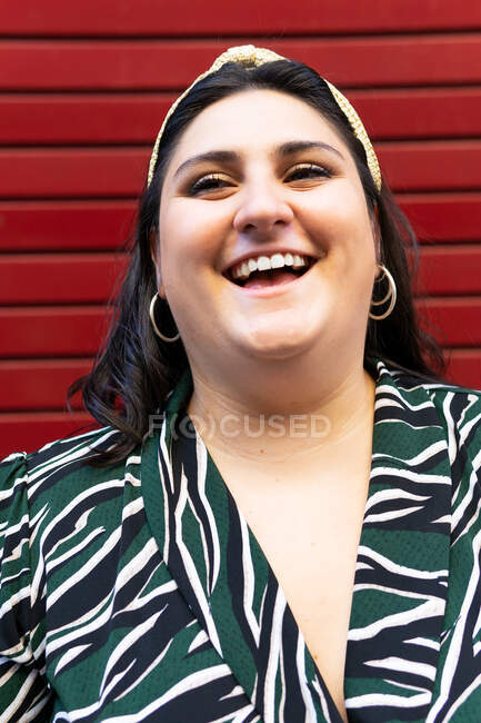 Portrait of cheerful young curvy brunette in stylish striped outfit and headband while looking at camera against red wall — Stock Photo
