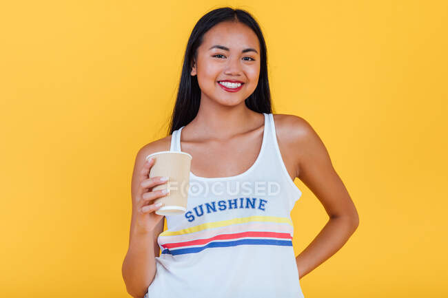 Delighted Asian female in summer outfit standing with takeaway cup of coffee on yellow background and looking at camera — Stock Photo