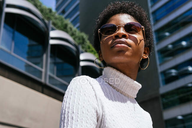 From below positive young African American woman in trendy outfit looking away and enjoying sunlight while sitting on bench on modern city street — Stock Photo