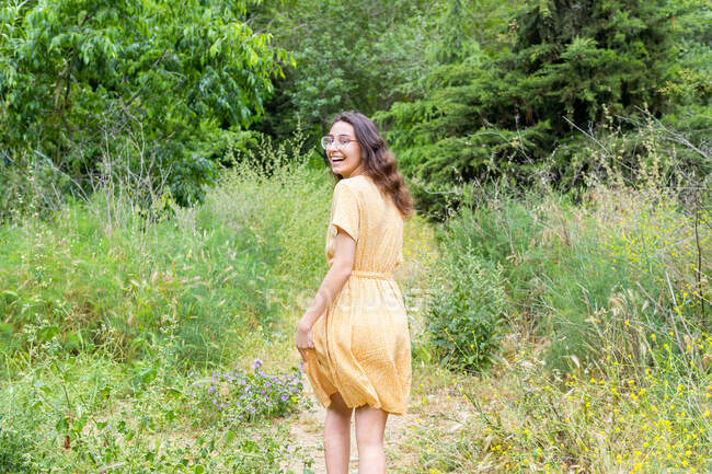 Vista posterior de la mujer encantada en vestido caminando en el parque y mirando a la cámara sobre el hombro en verano - foto de stock
