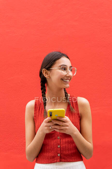 Веселая молодая женщина в косичках прическа просматривает на смартфоне стоя глядя в сторону на красном фоне на улице — стоковое фото