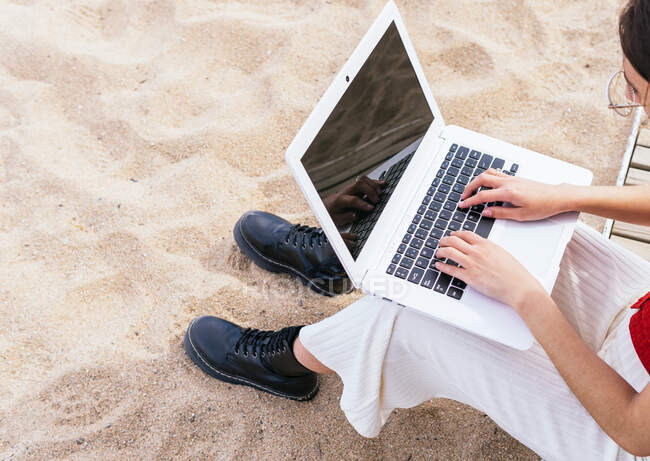 Desde arriba recortado irreconocible freelancer femenina sentada en la orilla del mar y escribiendo en el ordenador portátil mientras trabaja de forma remota en el proyecto en verano - foto de stock