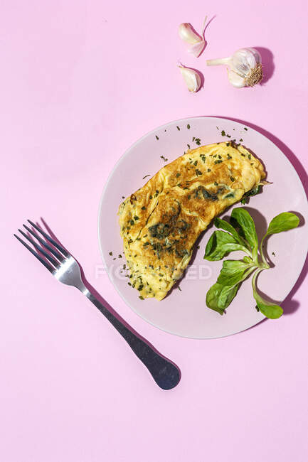 Tasty omelette on plate against fresh parsley sprigs with garlic cloves on pink background — Stock Photo
