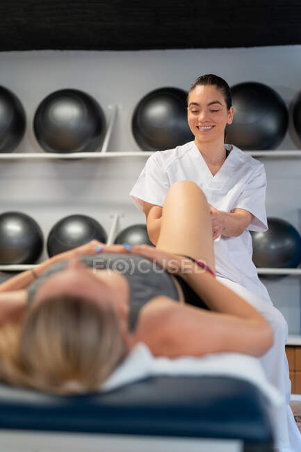 Happy masseuse in white robe massaging calf of anonymous female patient during physiotherapy session in clinic — Stock Photo