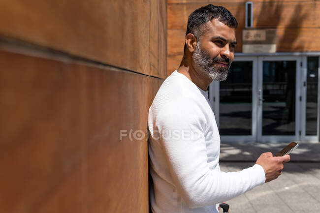 Vista laterale felice maschio barbuto in maglione bianco in piedi vicino muro edificio moderno e guardando altrove nei pensieri nella giornata di sole — Foto stock