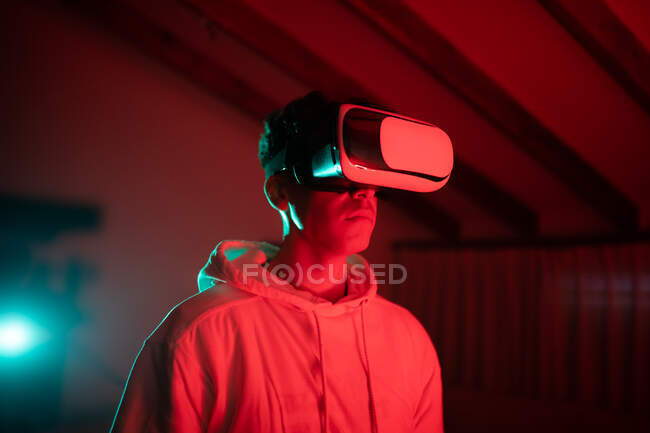 Homem usando capuz enquanto usa óculos VR e de pé no estúdio com luzes de néon vermelho — Fotografia de Stock