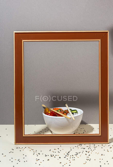 Hohe Winkel der weißen Schüssel mit leckeren Poke-Gericht und Essstäbchen hinter Rahmen auf dem Tisch mit Sesam bedeckt platziert — Stockfoto