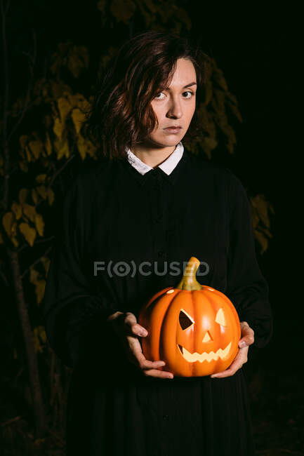 Donna in abito nero in piedi con lanterna di zucca incandescente in boschi scuri su Halloween e guardando la fotocamera — Foto stock