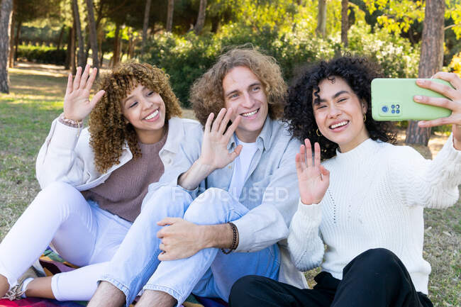 Group of young multiracial women and man with curly hair sitting on green grass in park and waving hands while taking selfie with mobile phone — Stock Photo