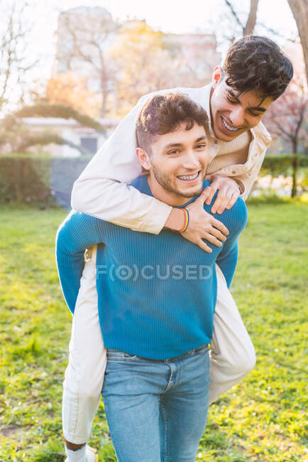 Positive gay Männchen huckepack lächelnden Freund, während Spaß zusammen im Park am Wochenende — Stockfoto