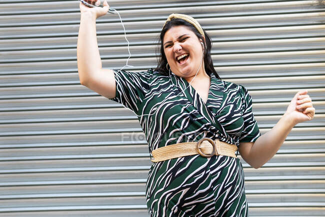 Happy young curvy female in stylish ornamental dress and headband listening to audio message via mobile phone with earphones and laughing while standing against wall of modern building — Stock Photo