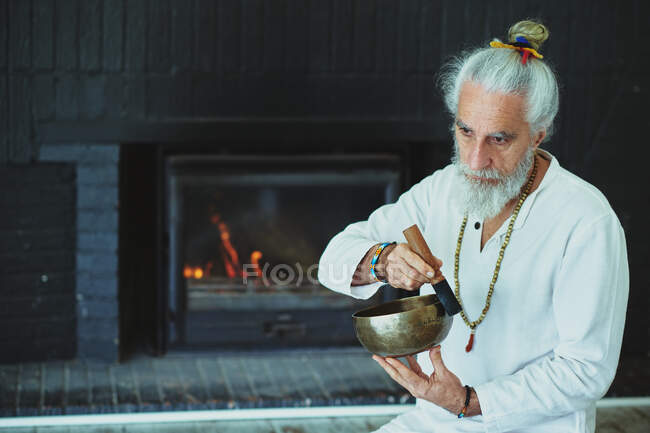 Elderly male with gray beard playing singing bowl with wooden striker while looking away during spiritual practice — Stock Photo