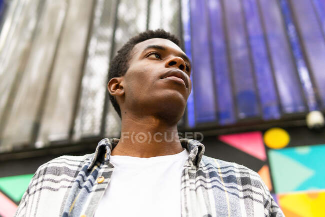 From below serious dreamy African American male standing looking away colorful background in the street — Stock Photo
