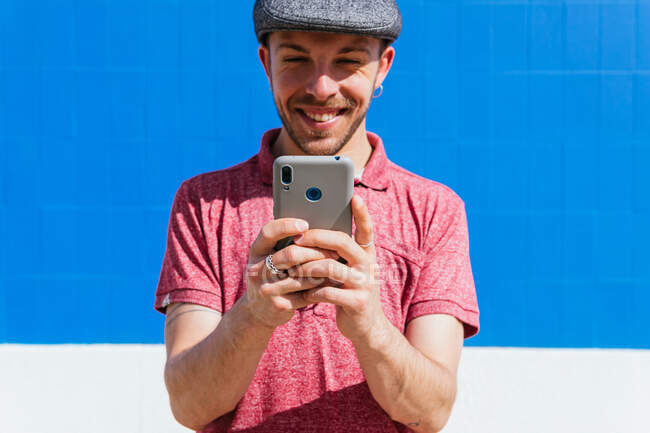 Зміст молодого бородатого хіпстера в повсякденній сорочці поло і кепці перегляду мобільного телефону, стоячи на синій стіні на сонячному світлі — стокове фото