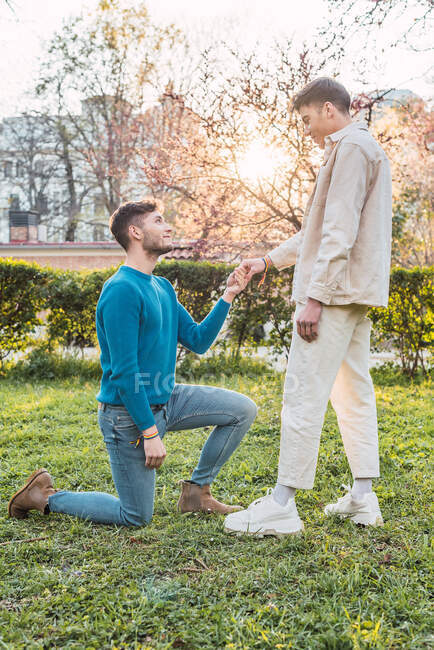 Vista lateral de homossexual masculino em pé no joelho e segurando a mão do namorado ao fazer proposta no parque — Fotografia de Stock