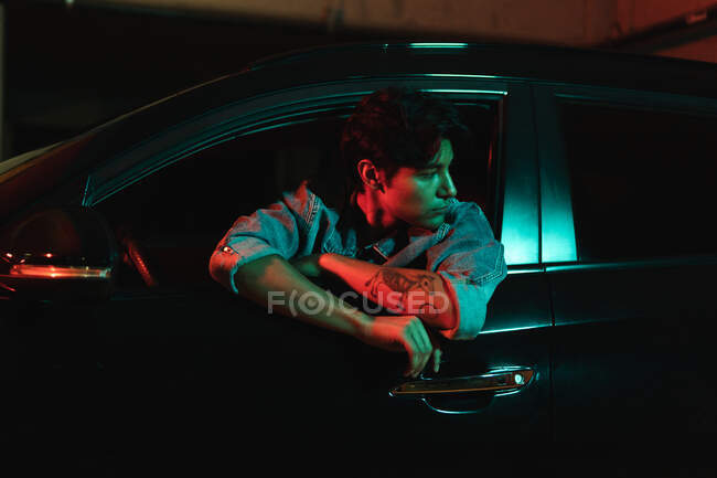 Young latin man sitting in car and looking away from driving seat under color lights at night — Stock Photo