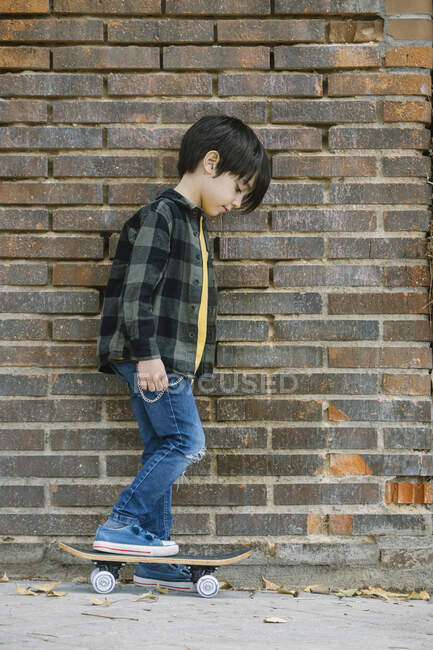Vue de côté corps complet de garçon hispanique ethnique en vêtements décontractés regardant vers le bas tout en se tenant sur skateboard au mur de briques sur la rue — Photo de stock
