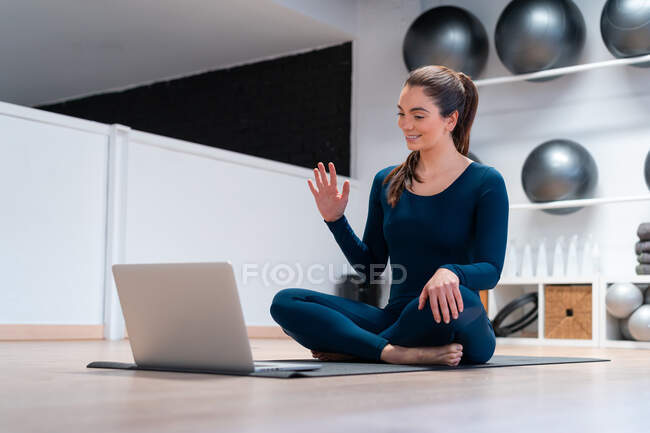 Ganzkörper positiver junger weiblicher Instruktor winkt mit der Hand, während er die Schüler während des Online-Yoga-Kurses per Videochat auf dem Laptop begrüßt — Stockfoto