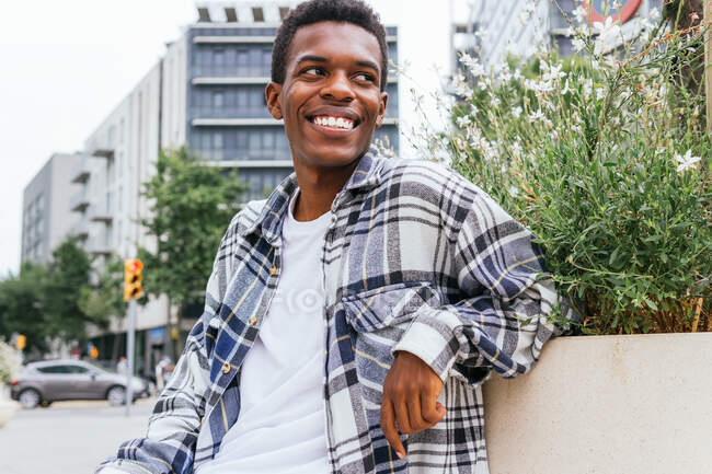 Homme élégant afro-américain joyeux assis sur un banc dans la ville et regardant ailleurs — Photo de stock
