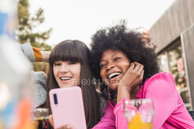 Pareja multiracial de mujeres homosexuales tomando uno mismo disparo en el teléfono móvil sentado en la mesa con cóctel en la cafetería de la calle - foto de stock