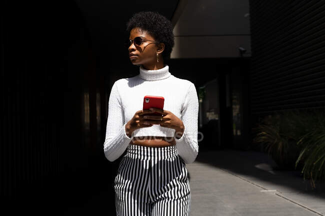 Afroamerikanerin in trendiger Kleidung surft auf ihrem Handy in den sozialen Medien, während sie an sonnigen Tagen auf der Straße der Stadt spaziert — Stockfoto