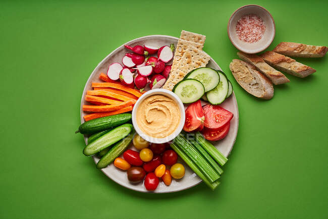 From above of appetizing fresh raw cut cucumbers with carrots and radish served on white table with assorted tomato and hummus and placed on green table with pieces of bread — Stock Photo