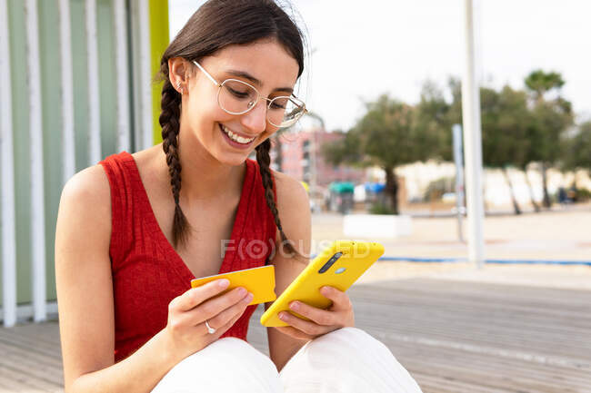 Contenu femelle payant pour commander avec une carte plastique pendant les achats en ligne via smartphone tout en étant assis dans la rue — Photo de stock