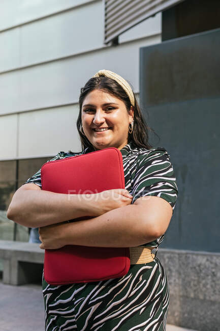 Весела молода жінка в стильному смугастому вбранні з червоним ноутбуком в руках посміхається і дивиться на камеру, стоячи проти сучасної міської будівлі — стокове фото