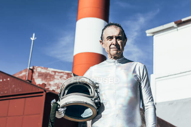 Low angle of senior male astronaut with helmet looking at camera while standing against shabby buildings of spaceport on sunny day — Stock Photo